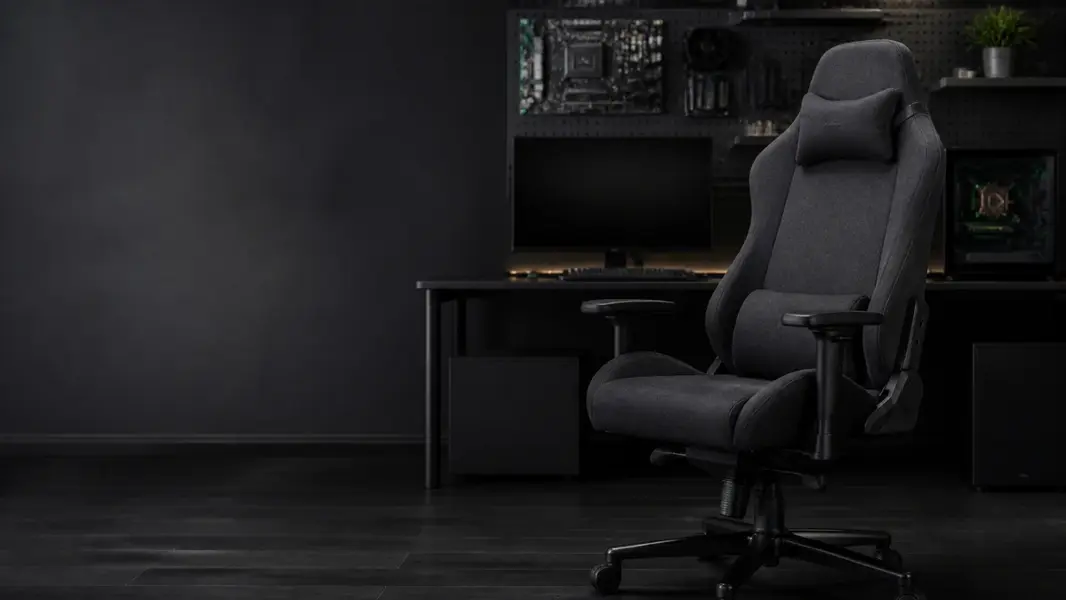 Mørkegrå gamerstol i stof med nakke- og lændepude foran et skrivebord med skærm og pc