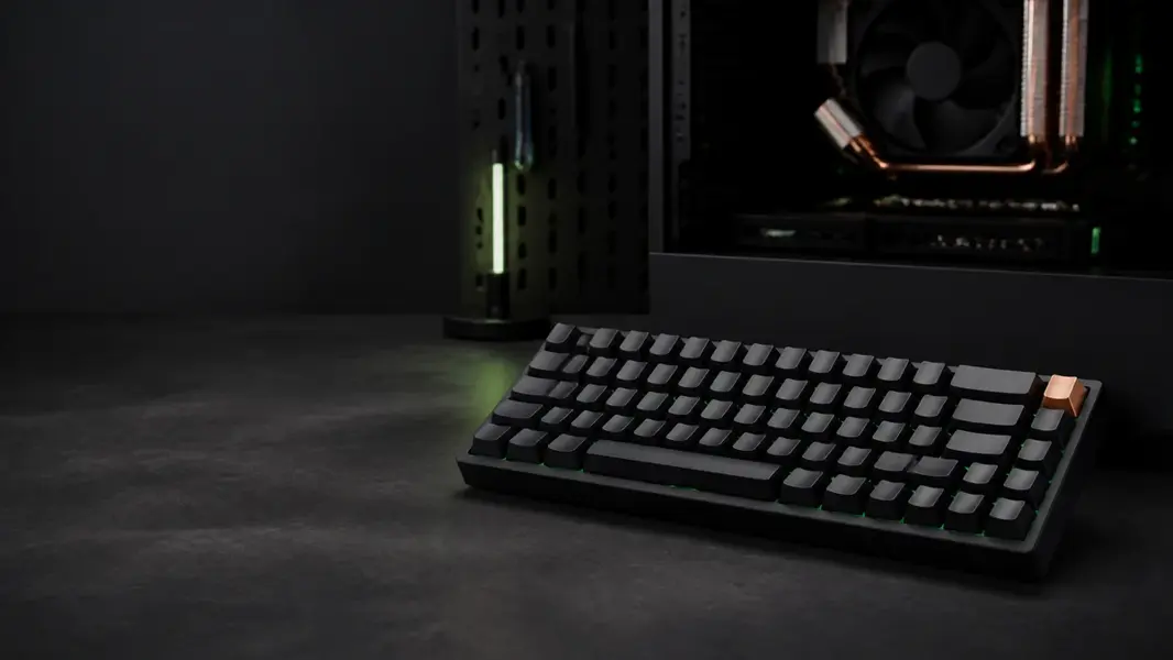 Kompakt sort mekanisk tastatur med en kobberfarvet tast foran et pc-kabinet med køler og grønt lys